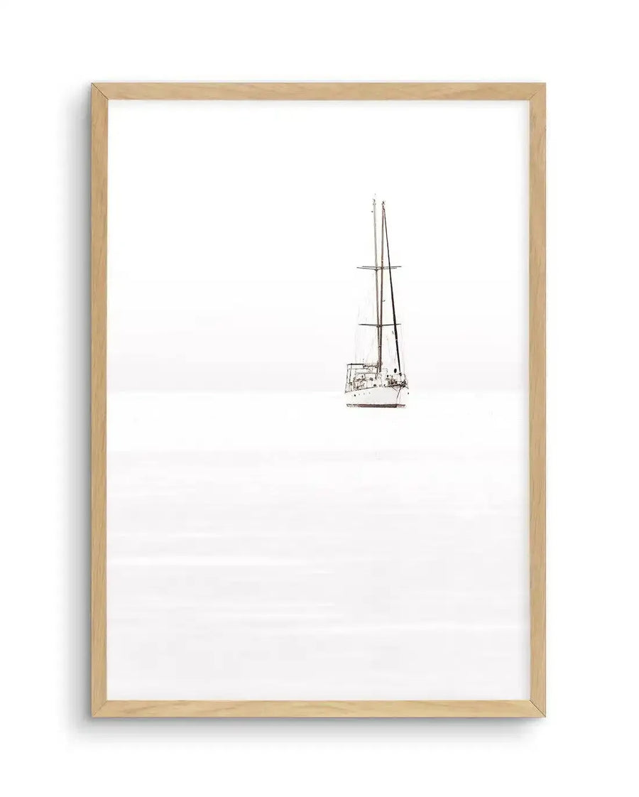 Framed black and white photograph of a sailboat on a vast, minimalist white background, displayed in a natural oak frame with white border. The sailboat, with its two tall masts and rigging, is positioned slightly to the right of the center, creating a sense of isolation and calm. The water below the boat is subtly textured with horizontal lines, suggesting gentle ripples or a hazy horizon.