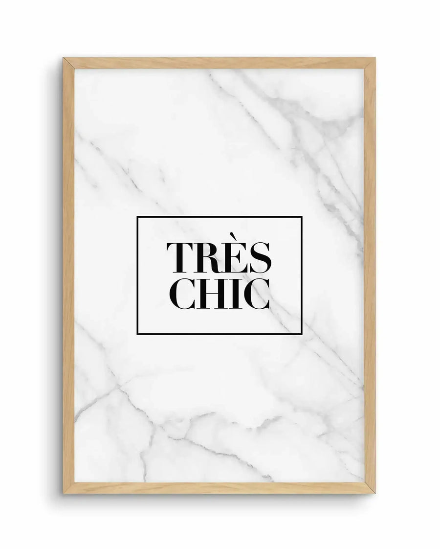 Framed art print with natural wood border displaying white marble-textured background with gray veining throughout. Black rectangular box centered on marble contains text reading "TRÈS CHIC" in large serif font with accent mark. White border surrounds the boxed text design. Wood frame has light natural finish against plain white background.