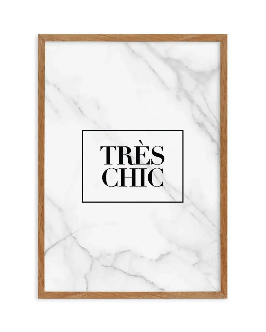 Framed art print featuring "TRÈS CHIC" in bold, serif black text centered within a black rectangular border on a marble-textured background. The background displays soft gray and white veining characteristic of marble. The print is mounted in a natural wood frame with a warm honey tone.