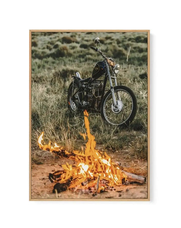 Framed canvas art print featuring a color photograph of a vintage motorcycle with black and chrome detailing, positioned in dry grassland with a bright campfire burning in the foreground. The motorcycle is shown in sharp focus against a softly blurred background of sage green vegetation and hills. Warm orange and yellow flames rise from the fire, creating a striking contrast with the earthy terrain and natural landscape. The canvas is mounted in a natural wood frame with a white border.