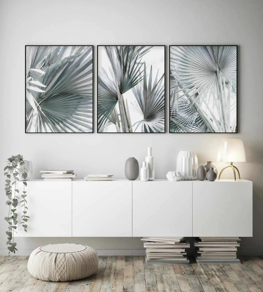 Three framed art prints displayed above a white console table in a minimalist living room. Each print features a close-up photograph of silver-green palm fronds against a bright white background, with a thin black frame and no border. The console table is adorned with various white and grey ceramic vases, a stack of books, and a gold-based table lamp with a white shade. A potted plant with trailing vines hangs from the left side of the table, and a round, cream-knitted pouf rests on the light wood floor.