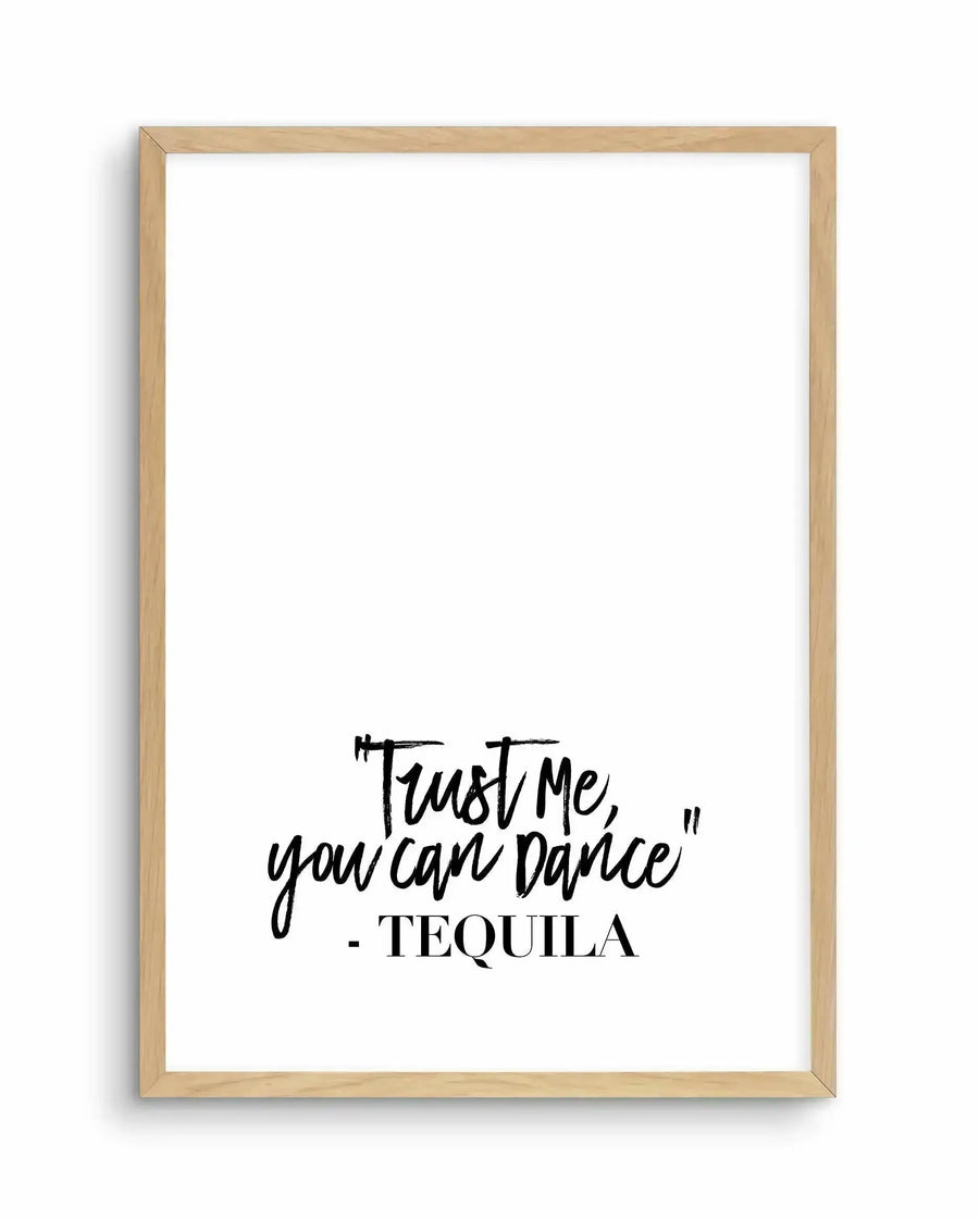 Framed art print with natural wood frame displaying a handwritten-style quote in black ink on white background. The text reads "Trust me, you can dance" in casual script lettering, followed by "- TEQUILA" in serif capital letters below. The composition is centered in the lower portion of the frame, leaving substantial white space above. The wood frame has a light natural finish with visible grain texture.