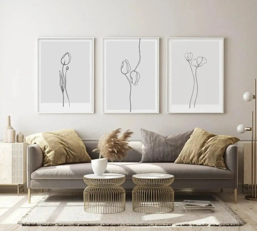 Lifestyle shot of three framed art prints displayed above a grey sofa in a modern living room. Each print features a minimalist black line illustration of tulips on a light grey background, with white frames and white borders. The sofa has gold and grey throw pillows, and two round gold side tables with white tops. A floor lamp with two white globe lights stands to the right.