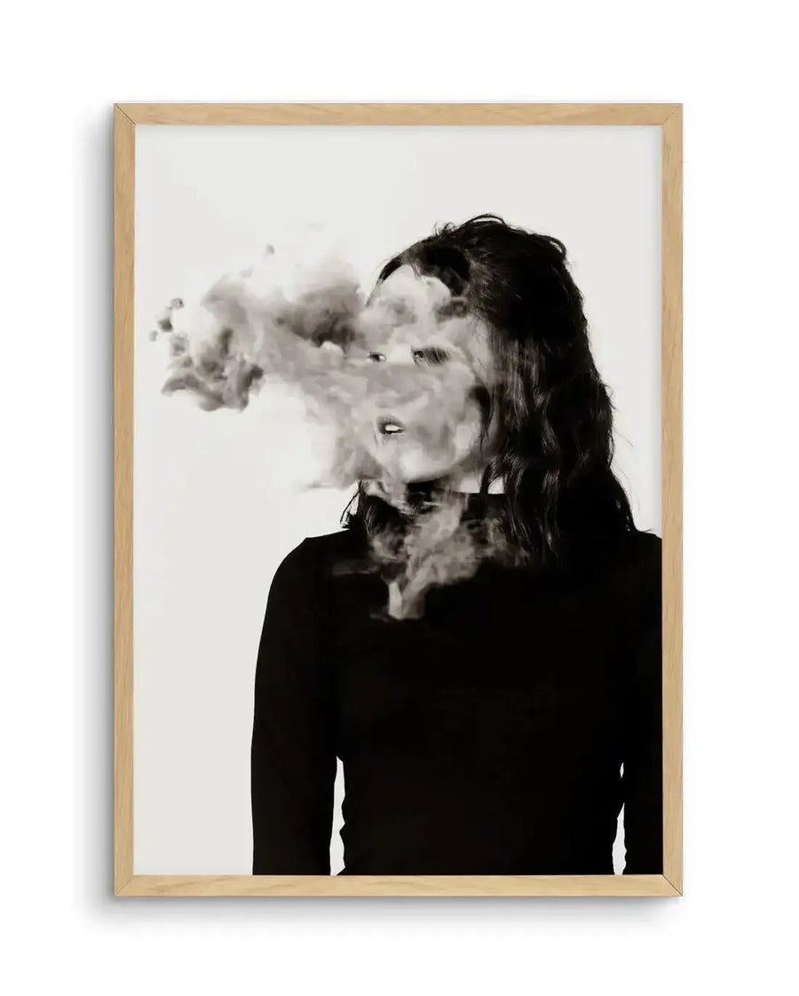 Framed black and white photography art print titled "Two Faced" featuring a woman with dark, wavy hair looking to the left, with a large plume of smoke obscuring her face and neck, creating a surreal and mysterious effect. The print is displayed in a natural oak frame with a white border.