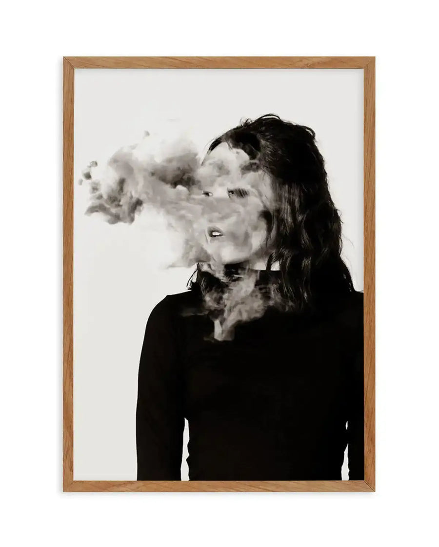 Framed black and white photography art print of a woman's profile with her face obscured by a large cloud of smoke. The woman has long, dark, wavy hair and is wearing a dark long-sleeved top, with a natural oak frame and white border.