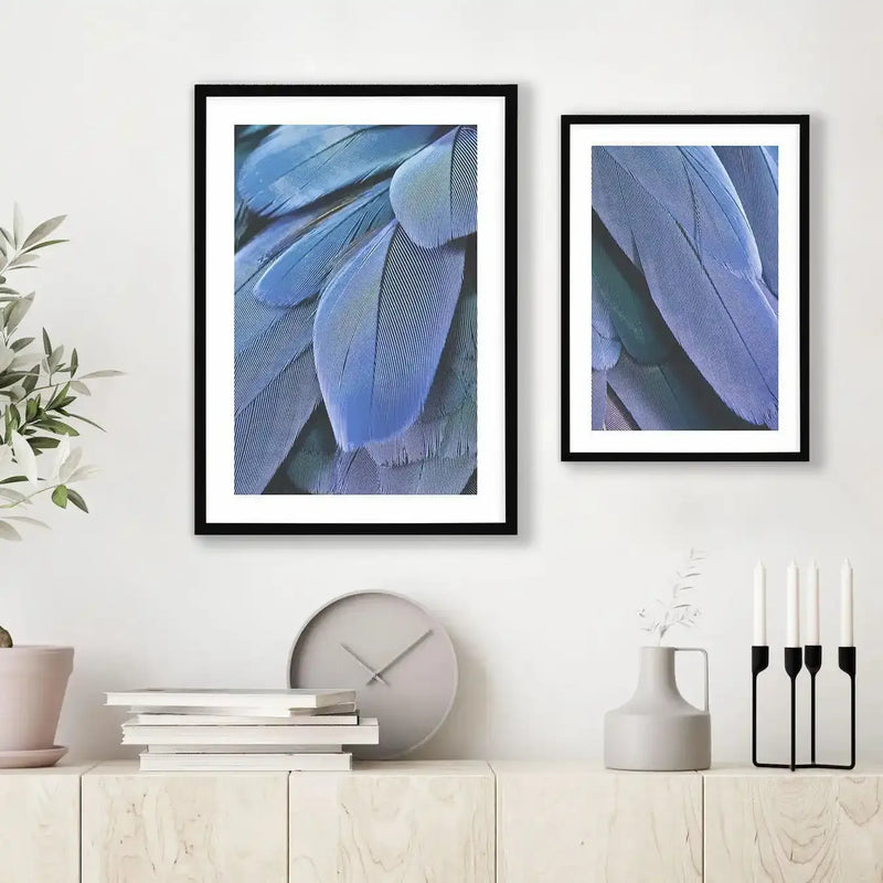 Lifestyle shot featuring two framed art prints displayed above a light wood cabinet in a minimalist room. The larger print on the left and smaller print on the right both feature close-up photographs of vibrant blue and purple bird feathers with fine, parallel lines of texture, displayed in black frames with white borders. The cabinet holds a stack of white books, a grey clock, a grey vase with a white branch, and a black candle holder with four white candles.