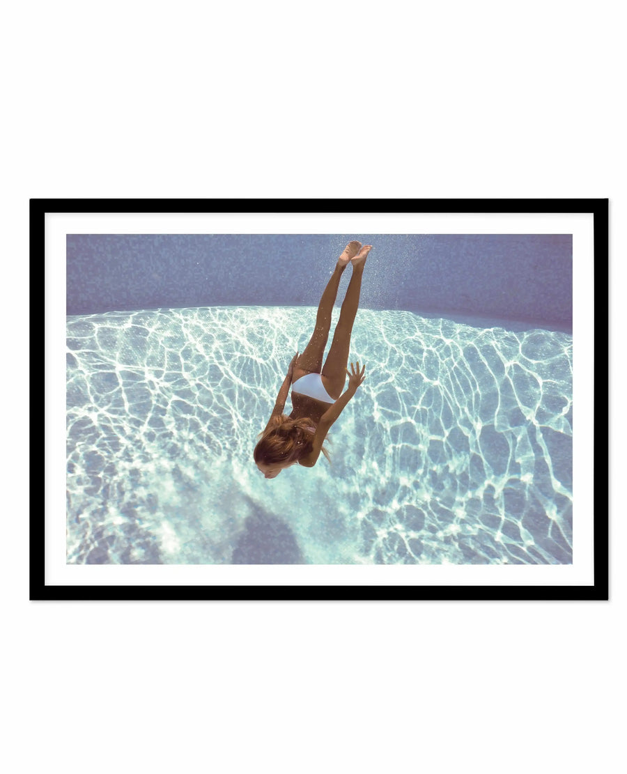 Framed art print featuring an overhead underwater photograph of a woman in a white bikini diving into crystalline turquoise water, viewed from above. Her body is captured mid-dive with arms extended upward and reddish-brown hair flowing. The water below displays rippling light patterns in shades of aqua, mint green, and pale blue. The sky above transitions from soft purple to deeper blue tones. Black frame with white border around the photographic image.