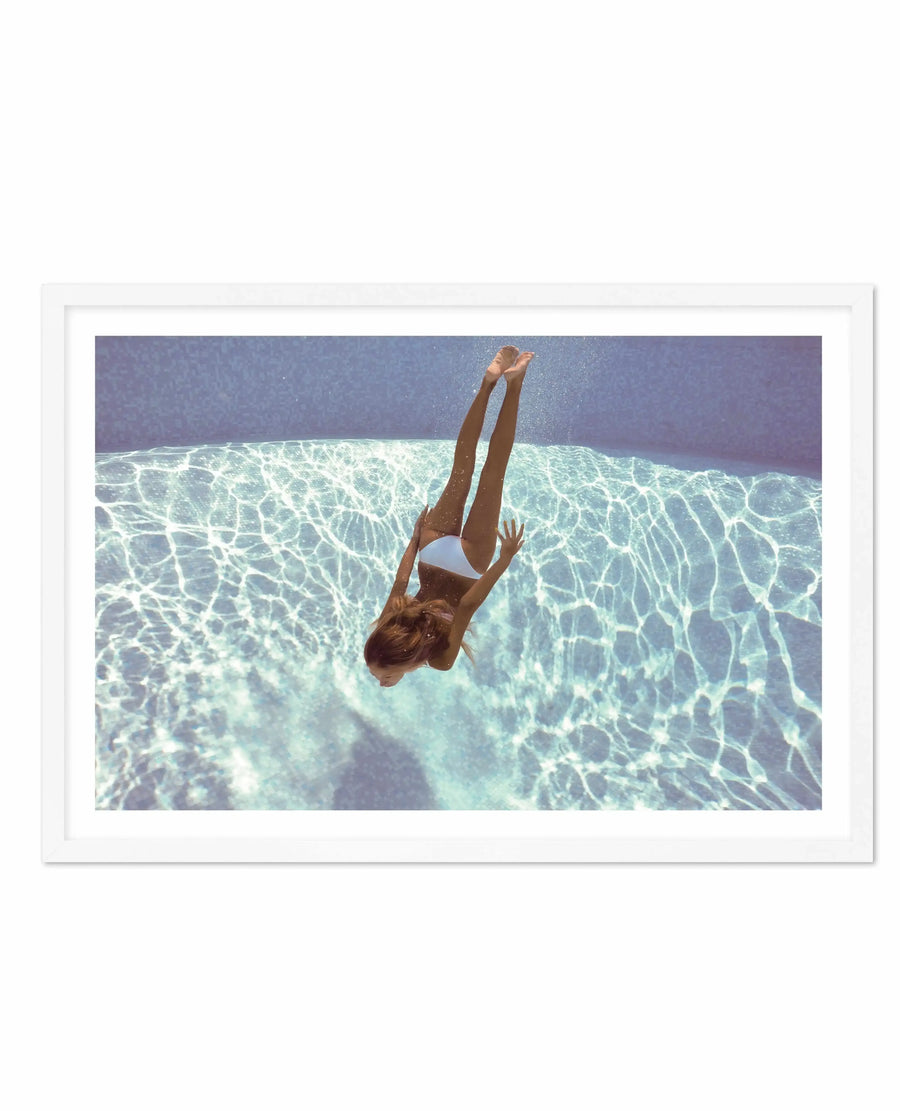 Framed art print featuring an overhead underwater photograph of a woman diving into clear turquoise water. Her body is captured mid-dive with arms extended upward and legs together, wearing a white bikini. The water displays rippling light patterns in shades of aqua, mint green, and cream, with darker blue-grey tones visible deeper below. The photograph has a dreamy, vintage quality with warm skin tones contrasting against the cool water palette. White wooden frame with white border.