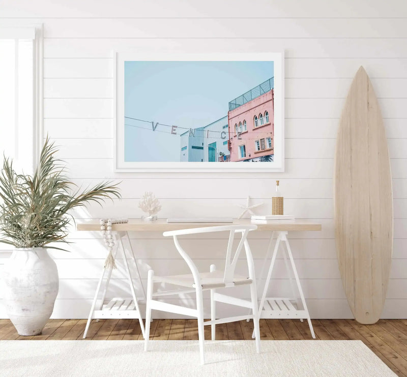 Framed art print displayed on white shiplap wall above light wood desk with white chairs. The print shows a photograph of a dusty rose pink building with turquoise doors and arched windows against clear blue sky, with clothesline and palm tree visible. White frame with white border. Natural light floods the minimalist coastal-themed workspace decorated with white ceramic vase, dried pampas grass, wooden surfboard, and neutral decor.