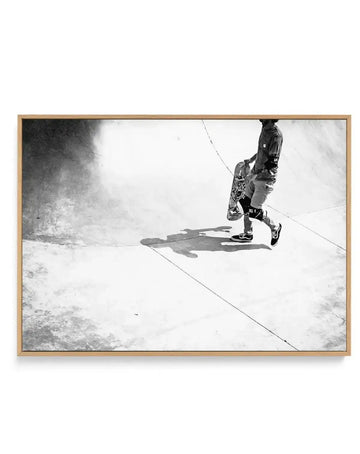 Black and white photograph of a skateboarder captured from above at Venice Skate Park, showing the athlete mid-trick on a concrete surface with their shadow visible below. The image is mounted in a natural wood frame with a white border, displayed as a horizontal canvas print against a white background.