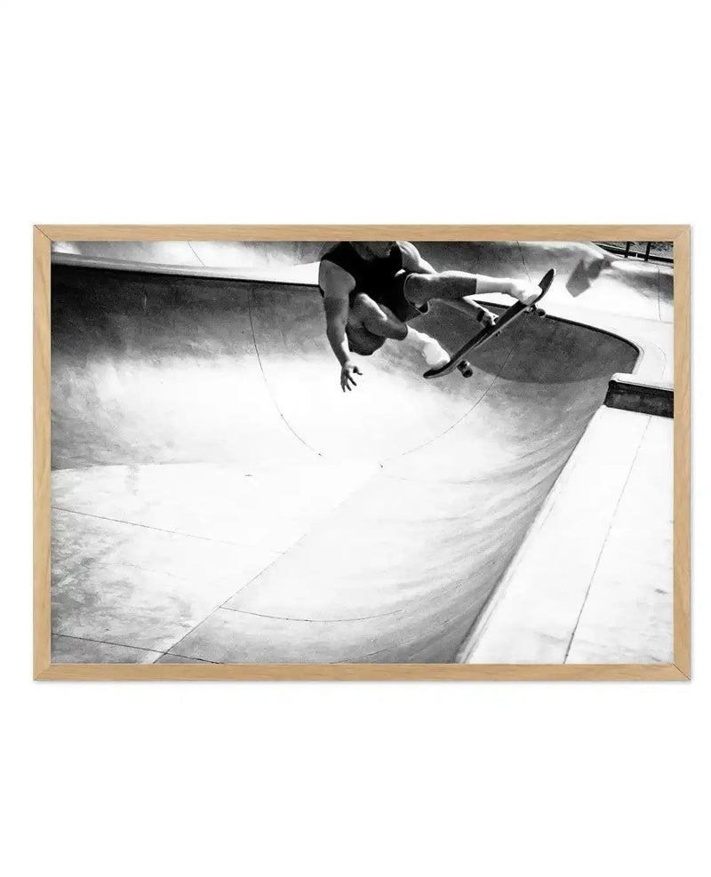 Framed black and white photograph of a skateboarder in mid-air performing a trick in a concrete skate park bowl, displayed in a natural oak frame with white border. The composition captures the dynamic movement of the skater, with the curved lines of the bowl creating a sense of speed and action.