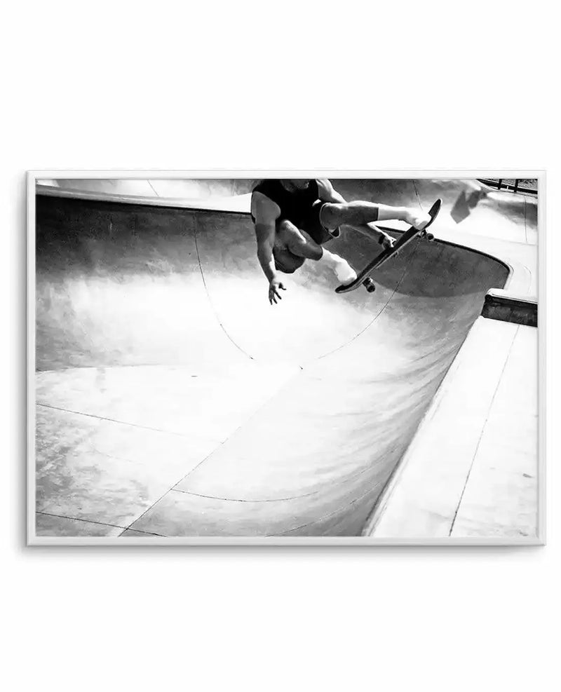 Framed black and white photograph of a skateboarder mid-air in a concrete skate park, with a white frame and white border. The composition captures the dynamic movement of the skateboarder in a tank top and shorts, suspended above the curved ramps and bowls of the park.