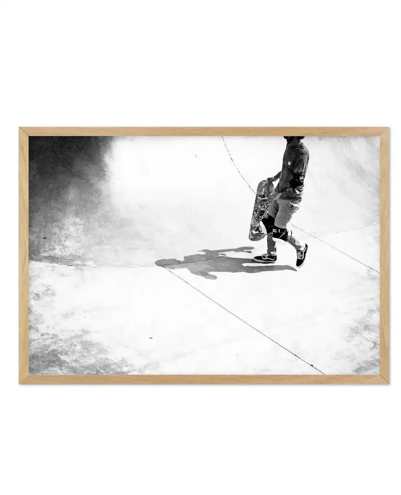 Framed black and white photograph of a person walking through a skate park, displayed in a natural oak frame with white border. The composition features a person in dark clothing and sneakers, holding a skateboard, casting a long shadow on the light concrete ground. The background shows the curved walls of the skate park with varying shades of grey and white.