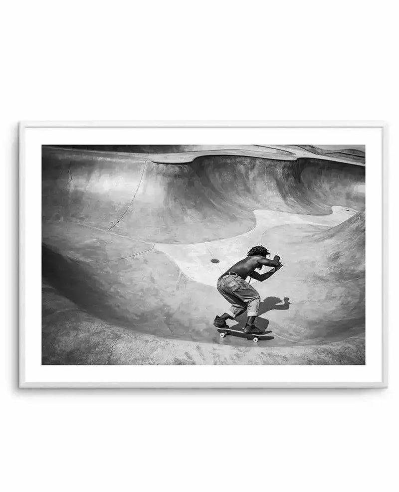 Black and white photograph of a shirtless skateboarder riding in a concrete skate bowl with curved, textured walls. The skater is captured mid-motion on the flat section of the bowl floor, leaning forward in an athletic stance. Dramatic shadows and light create depth across the weathered concrete surfaces. The composition emphasizes the scale and architecture of the park. Framed in a light gray frame with white border.