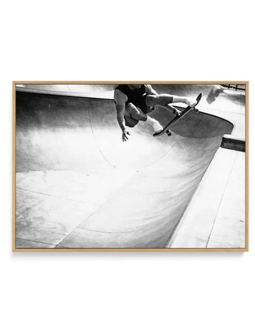 Black and white overhead photograph of a skateboarder riding down a curved concrete skate park bowl, captured from directly above. The skater is shirtless and mid-action on the smooth, curved concrete surface with dramatic shadows and light creating depth. The image is printed on canvas and mounted in a natural wood frame with a white border, emphasizing the documentary-style photography and architectural lines of the skate park bowl.