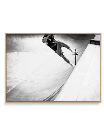 Black and white photograph of a skateboarder performing a trick in a concrete bowl at Venice Skate Park, captured from above at a dynamic angle. The skater is airborne against the curved concrete surface, wearing casual clothing and a cap. The composition emphasizes the smooth, weathered texture of the concrete with strong diagonal lines and dramatic lighting. Printed on canvas and framed in natural wood with clean edges.