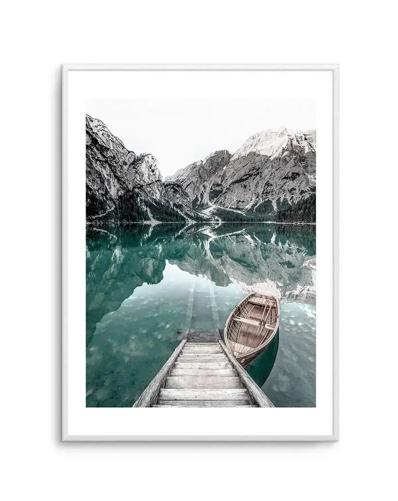 Framed photography art print featuring a serene mountain lake scene with a wooden boathouse and boat, displayed in a white frame with a white border. The composition captures towering, rugged mountains with snow-capped peaks reflecting in the calm, turquoise-green water. A wooden dock extends into the foreground, leading to a small, empty wooden rowboat gently floating on the water. The sky is bright white, and the overall mood is tranquil and expansive.