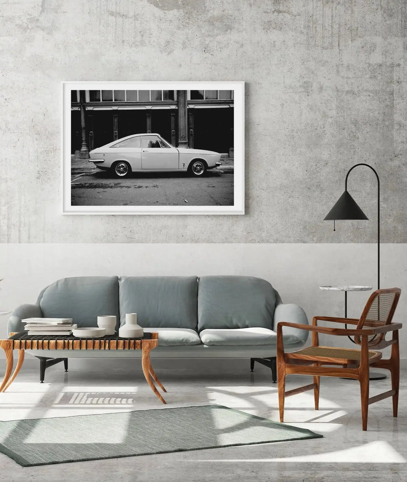 Black and white photograph of a classic 1970s Opel Manta GT coupe positioned in profile against an industrial brick building with large windows, printed and framed in white, displayed above a sage green curved sofa in a minimalist living room with mid-century wooden furniture and concrete walls.