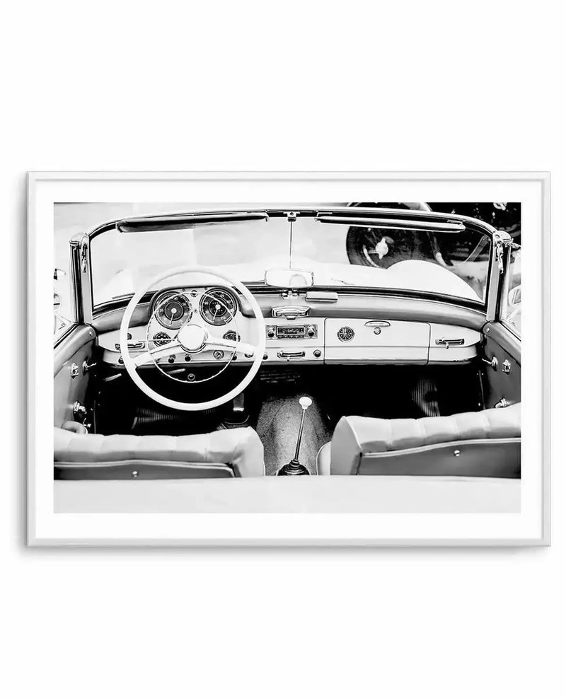 Black and white photograph of a convertible car interior from the 1950s, framed in silver aluminum with white border. The image shows a steering wheel with a light-colored rim, dashboard with vintage gauges and radio, manual gear shift, and light-colored upholstered seats. The windshield and convertible top frame are visible above. The photograph has high contrast and classic mid-century aesthetic. Print displayed in horizontal orientation within silver frame.