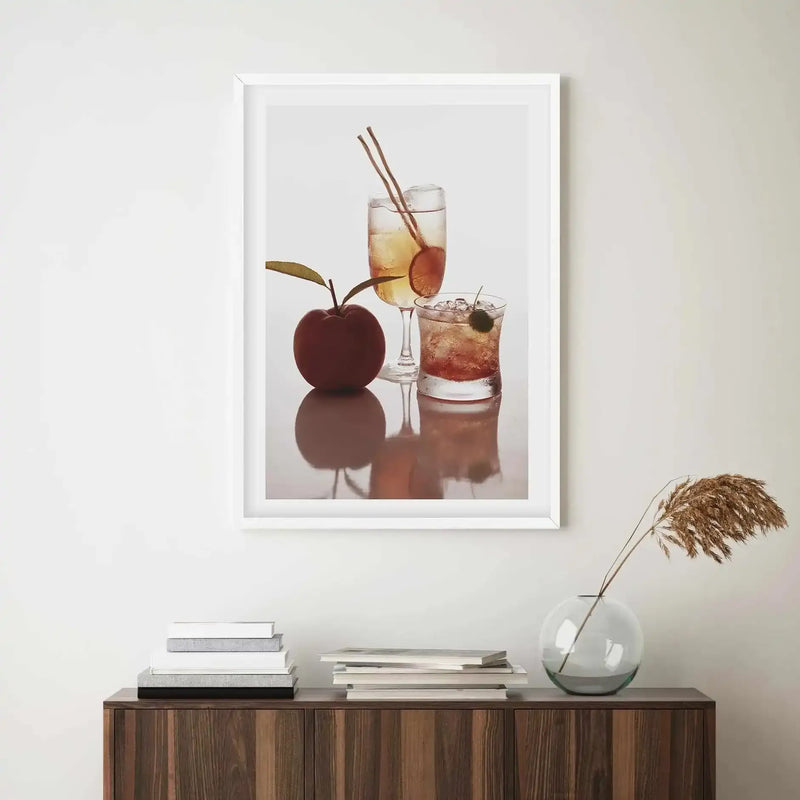 Framed art print displayed above a dark wood cabinet in a minimalist room. The print features a still life photograph of two cocktails and a red apple on a reflective surface against a white background. The taller cocktail is in a champagne flute with two cinnamon sticks and a citrus slice, while the shorter cocktail is in a tumbler with ice and a cherry. The room includes a stack of books and a clear glass vase with dried pampas grass.