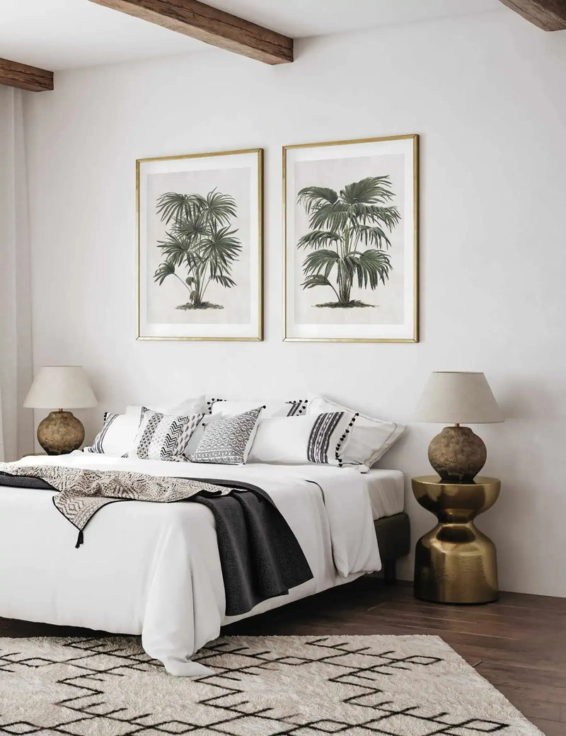 Two framed botanical art prints featuring detailed pen and ink illustrations of palm trees, displayed on a white bedroom wall above a white bed with patterned pillows and black throw blanket. Each print is set in a brass-toned frame with white border. Dark wooden ceiling beams, brass side tables with rounded ceramic lamps, and a geometric patterned area rug complete the room setting.