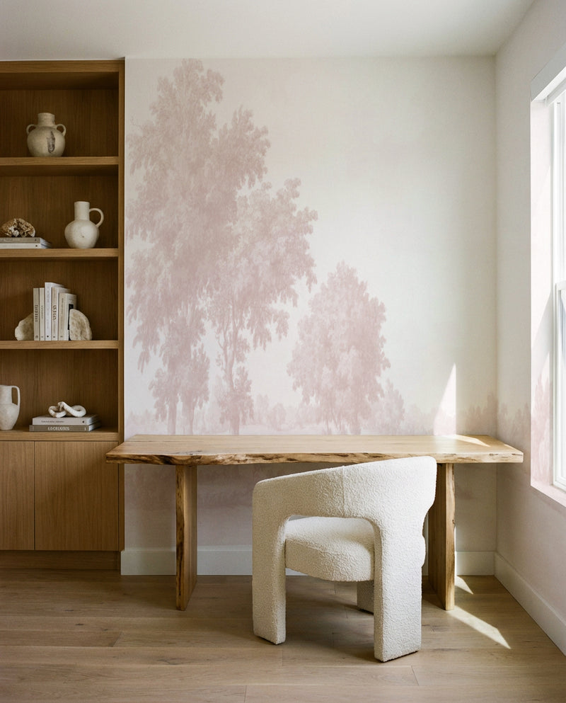 Dusty rose and blush pink watercolor illustration of tall evergreen trees rendered in soft, misty brushstrokes on cream-white wallpaper mural. Trees fade into the background creating depth. Mural installed on bedroom wall behind natural wood desk with live edge top and cream boucle curved chair. Adjacent wooden shelving unit with natural finish displays ceramic vessels, white books, and decorative objects. Bright natural light streams through window on right side casting soft shadows on wood flooring.