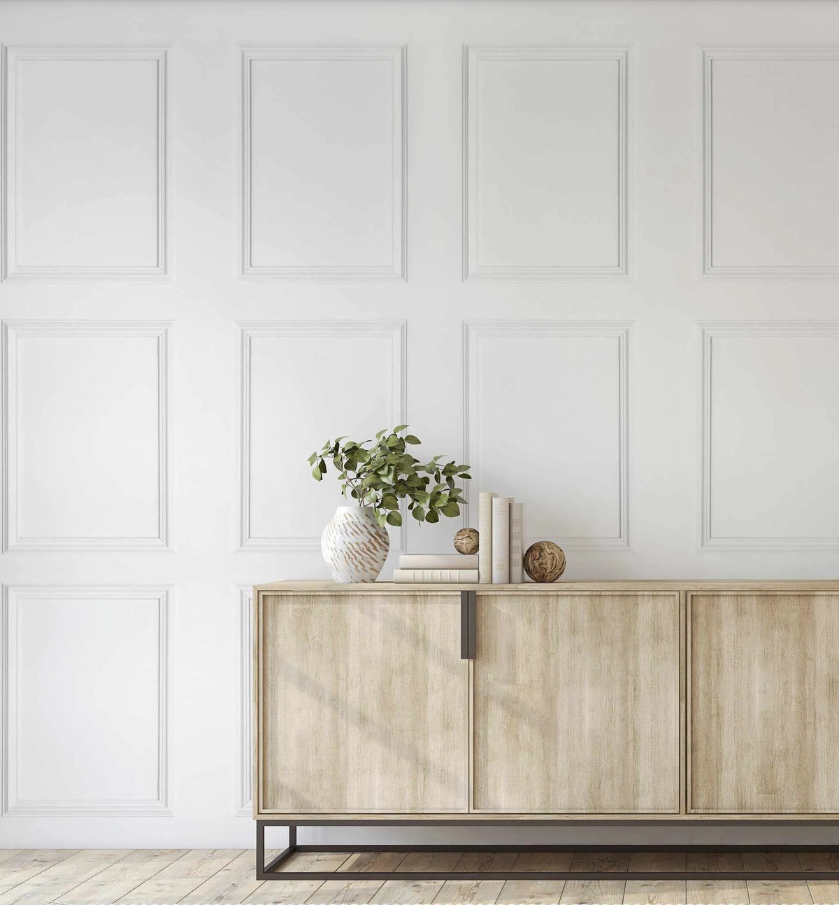 Lifestyle shot of a room featuring wainscoting Parisian panel wallpaper in a crisp white finish, creating a classic and elegant backdrop. The wallpaper features a repeating pattern of rectangular panels with raised, decorative trim. A light wood sideboard with dark metal legs is positioned against the wall, adorned with a white textured vase holding green leafy branches, a stack of white books, and two decorative wooden spheres.