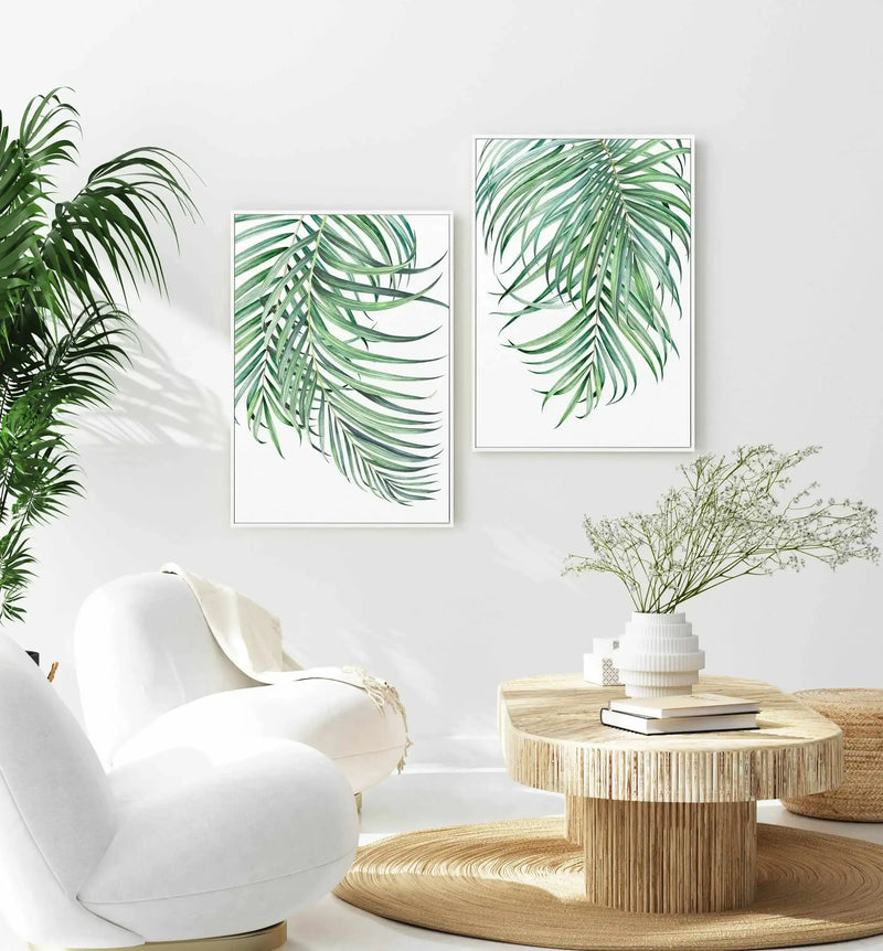 Two framed canvas art prints displayed side-by-side on a white wall in a bright, minimalist living room. Each print features a watercolour illustration of a palm frond in shades of emerald and sage green against a white background, with a thin white frame. A large green plant is visible on the left, casting shadows on the wall. A white armchair is in the foreground, and a round wooden coffee table with a white vase of white flowers sits on a woven rug.