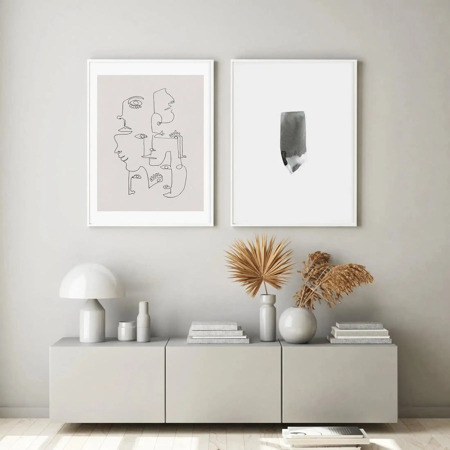 Abstract black watercolour stroke on white background, displayed as framed art print with white frame and white border, hanging on neutral beige wall above white storage unit styled with ceramic vessels, dried flowers, stacked books, and decorative objects.