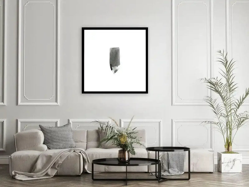 Watercolour Stroke Black Square Art Print with black frame displayed on a light grey panelled wall above a cream upholstered sofa. The artwork features a single grey-green watercolour brushstroke on a white background, positioned centrally within the black square frame. The print is shown in a modern living room setting with a black side table, potted pampas grass, and houndstooth patterned cushions.