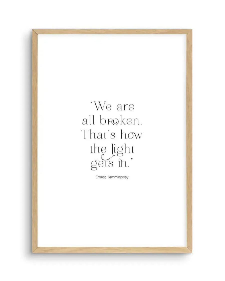 Art print featuring a quote in black serif font on a plain white background, displayed in a natural oak frame with white border. The quote reads: "We are all broken. That's how the light gets in." followed by "Ernest Hemingway".