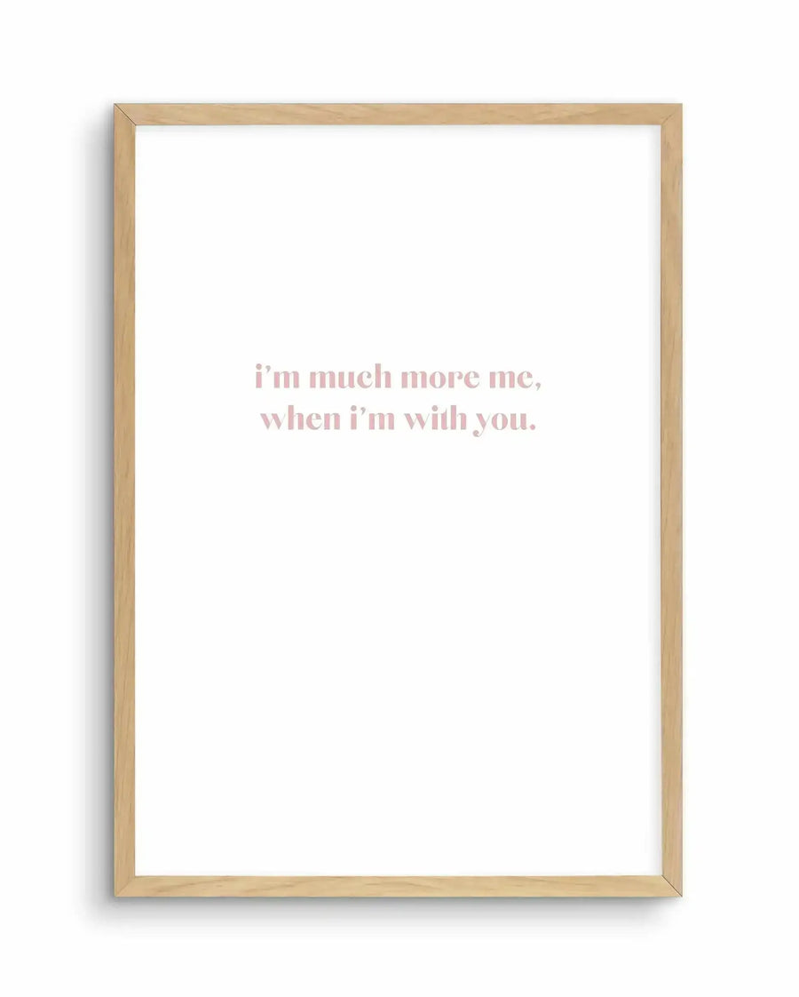 Framed art print with natural wood frame displaying text in dusty rose pink lettering centered on a white background. The text reads "i'm much more me, when i'm with you." in an elegant serif typeface. The wooden frame has a light natural finish and appears to be hung on a white wall, casting subtle shadows that indicate dimension and depth.