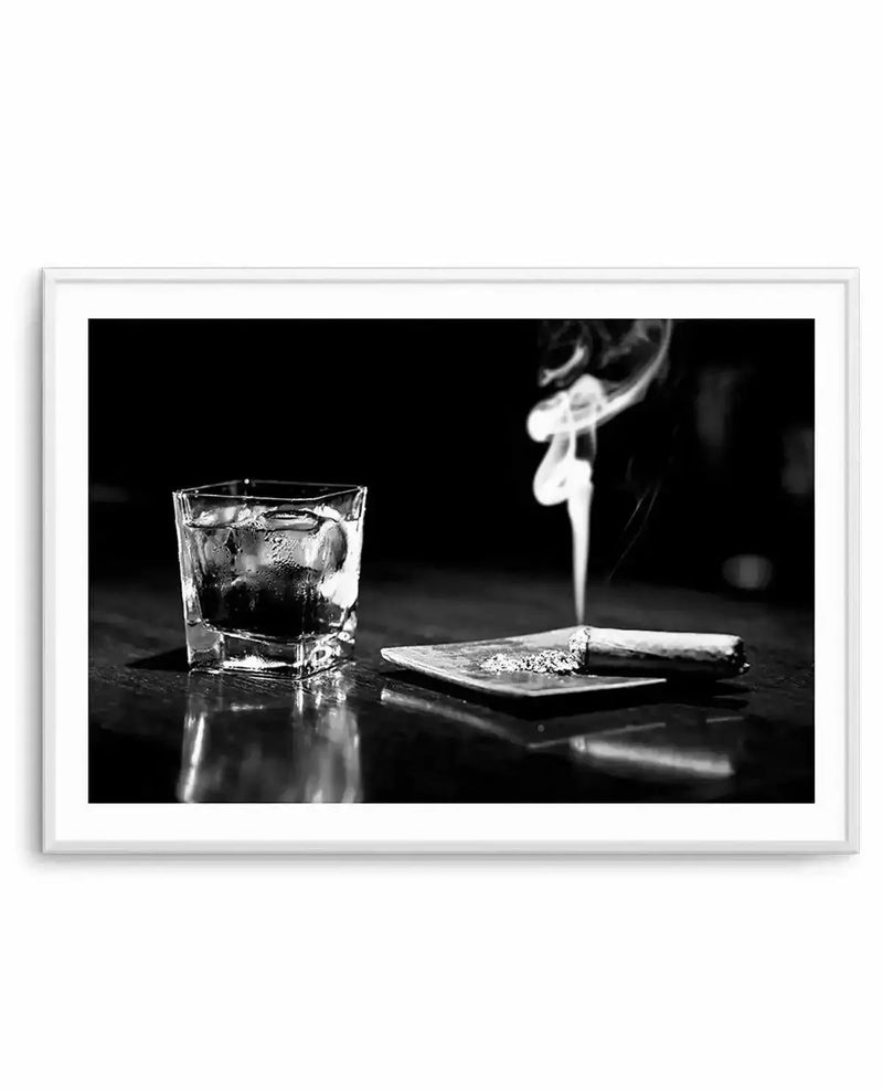 Framed black and white photograph of a glass of whisky with ice and a lit cigar on an ashtray, displayed in a white frame with white border. The composition features a square glass of whisky with condensation on the left, and a smoking cigar with ash on a small square ashtray on the right. The items are placed on a reflective surface, possibly a dark wooden table, with a dark, out-of-focus background. The smoke from the cigar rises in elegant curls.