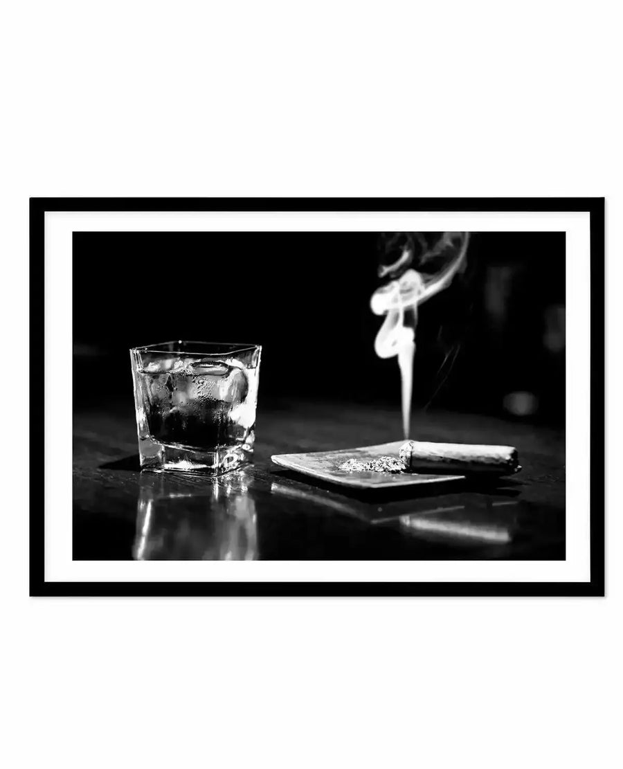 Black and white framed art print featuring a moody still life composition. A rocks glass filled with dark liquid and ice sits on a reflective black surface alongside a small plate with a lit cigar and scattered tobacco. Smoke rises from the cigar in the upper right. The photograph has high contrast with dramatic lighting that creates reflections on the dark surface. The image is mounted in a black frame with a white border.