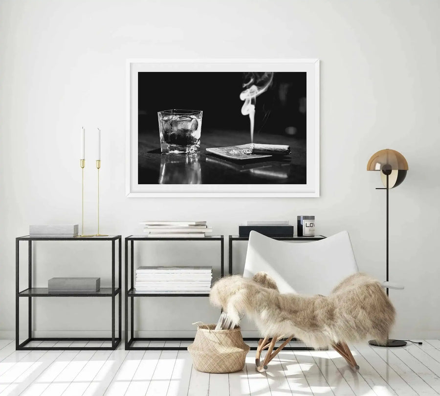 Black and white fine art photograph displayed in a white frame with white border, showing a glass of whisky with ice on the left and a lit cigar on a dark surface on the right, with dramatic lighting and reflections. The photograph is hung on a light gray wall above dark metal shelving units styled with books, candles, decorative objects, a cream-colored chair with faux fur throw, and a brass floor lamp.