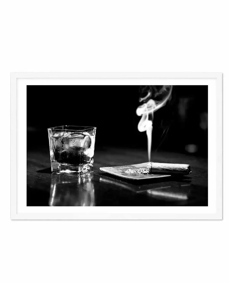 Black and white photograph in white frame with white border depicting a moody still life composition. A crystal glass filled with dark liquid and ice sits on a reflective dark surface alongside a small plate containing a lit cigar with visible ash and smoke rising upward. The dramatic lighting creates strong contrasts and highlights the condensation on the glass and reflective qualities of the surface, evoking a sophisticated, contemplative atmosphere.