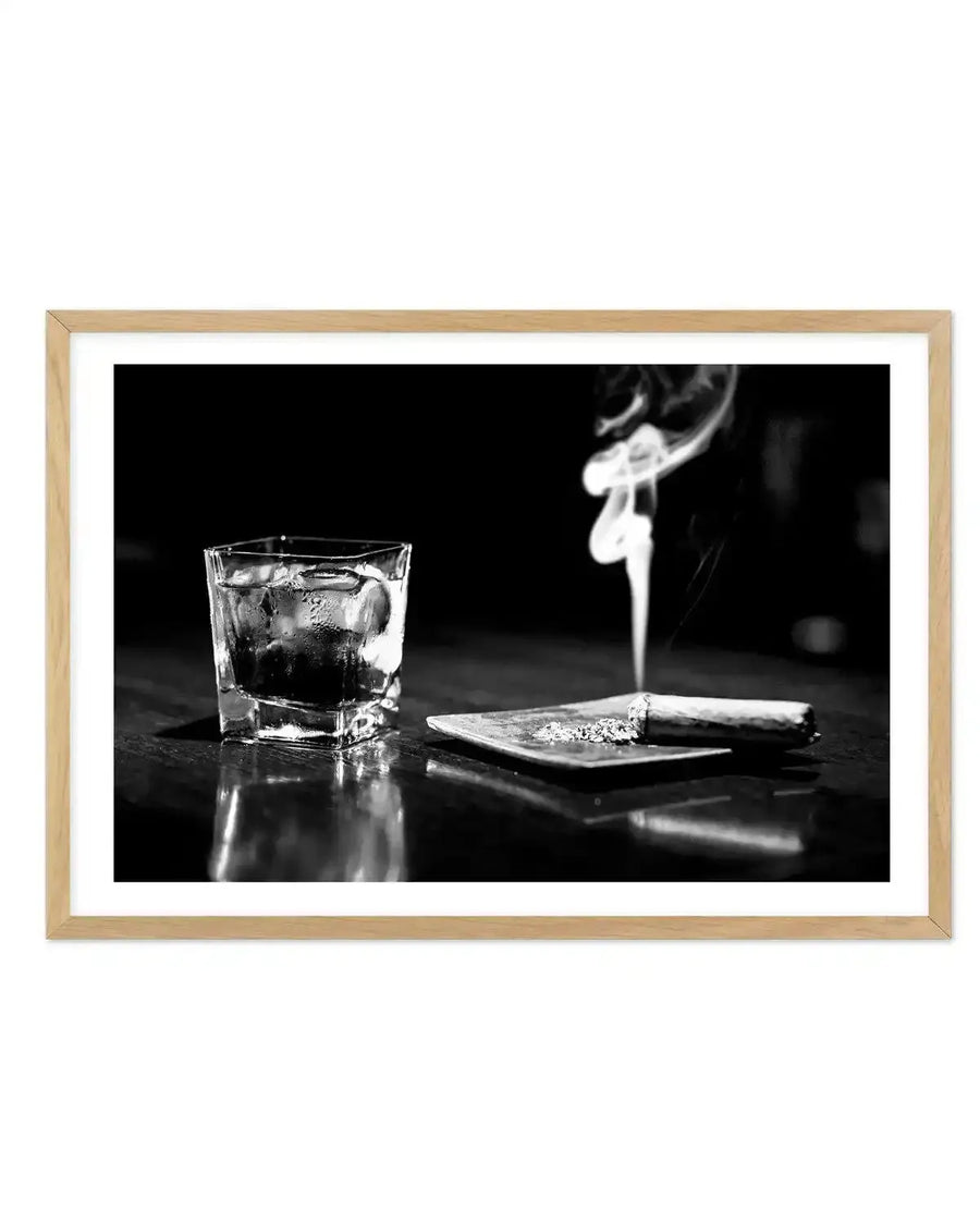 Black and white photograph printed on white paper with natural wood frame. The image shows a glass tumbler filled with dark liquid and ice on the left, positioned next to a small rectangular dish containing a lit cigar with ash, set against a dark reflective surface. Smoke rises from the cigar in the background. The photograph has high contrast with dramatic lighting that emphasizes the glass's transparency and the surface reflections. The composition conveys a sophisticated, moody aesthetic.