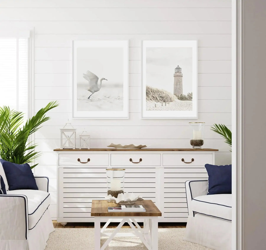 Lifestyle shot of a coastal-themed living room featuring two framed art prints above a white console table. The left print is a black and white photograph of a white bird in flight, captured mid-stride in shallow water. The right print is a black and white photograph of a lighthouse on a sandy beach with tall grasses. Both prints have white frames and white borders. The room has white shiplap walls, white armchairs with navy blue piping and cushions, a wooden coffee table, and green potted plants.