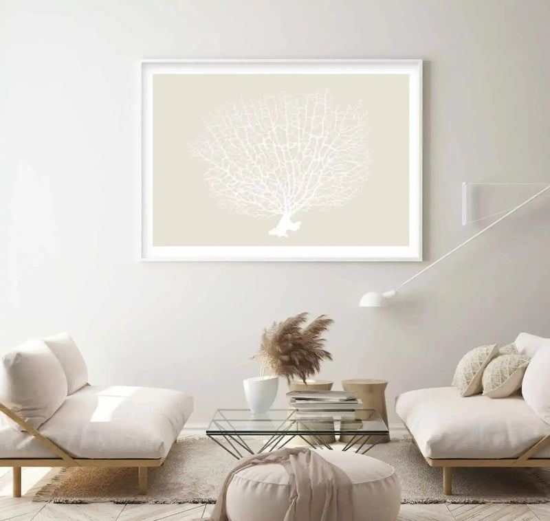 Large framed art print displayed above two cream sofas in a minimalist living room. The print features a white coral on a light beige background, with a white frame and white border. The room includes a glass coffee table with books and a vase of dried pampas grass, a cream ottoman, and a white floor lamp.