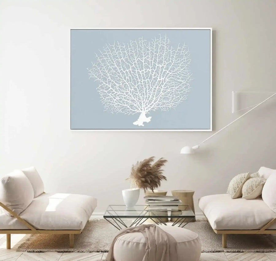Framed canvas art print displaying a white silhouette of branching coral against a soft dusty blue background, hung on a neutral beige wall above two cream-colored cushioned seating pieces flanking a glass coffee table in a minimalist living room setting. The canvas features a thin gray frame with white border.