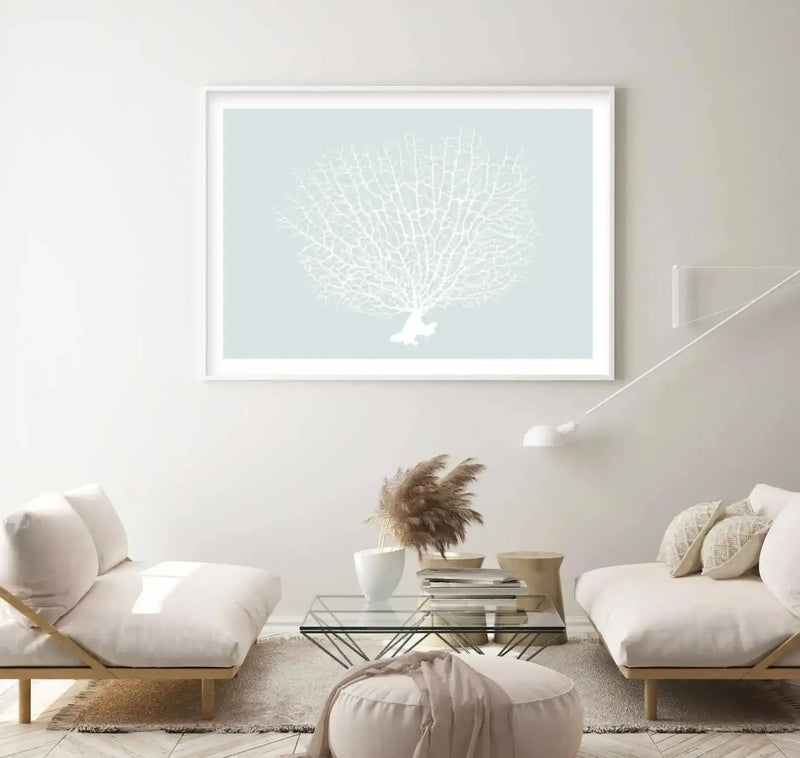 Large framed art print displayed above a glass coffee table in a minimalist living room. The print features a white coral fan with intricate branching details against a soft mint green background, displayed in a white frame with a white border. The room includes two cream-colored sofas, a white pouf, dried pampas grass in a white vase, and a modern white wall lamp.