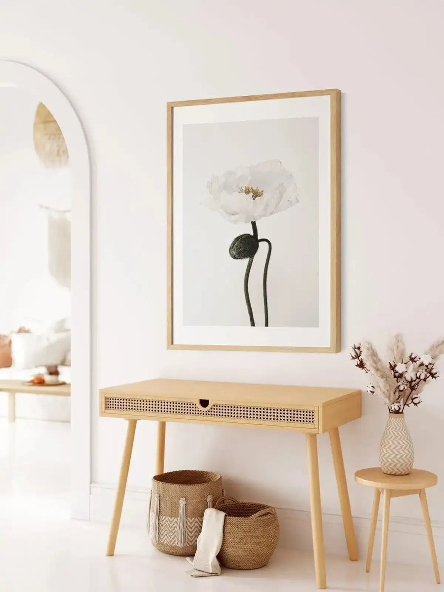 Lifestyle shot of a minimalist entryway featuring a framed art print of a white poppy above a light wood console table. The print depicts a single white poppy with delicate, ruffled petals and a dark green seed pod on a slender stem, set against a soft white background, displayed in a natural oak frame with a white border. The console table has a woven rattan drawer front and is accompanied by two woven baskets and a small round stool with a vase of dried pampas grass and cotton branches.