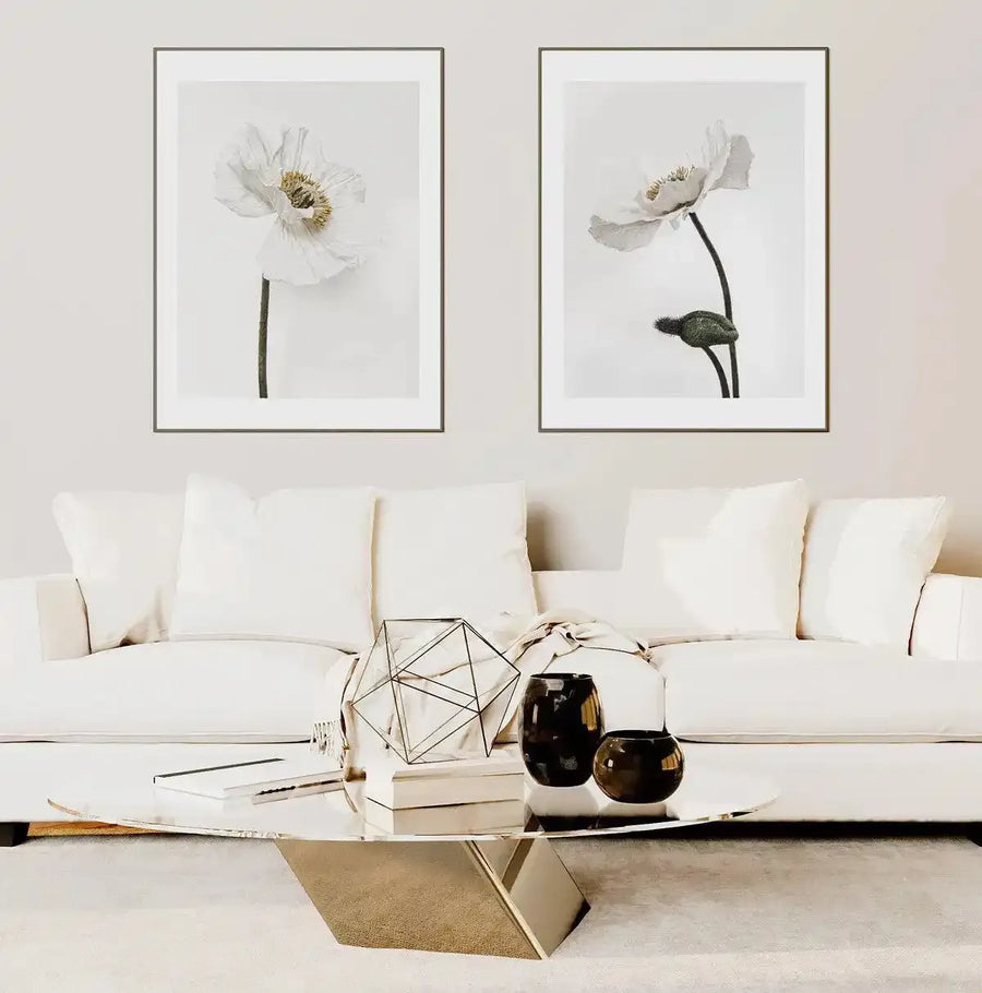 Lifestyle shot of two framed art prints displayed above a white sofa in a minimalist living room. The left print features a white poppy with a yellow center and green stem against a white background. The right print shows a white poppy with a green stem and closed bud against a white background. Both prints have a natural wood frame and white border. A metallic coffee table with books, a geometric sculpture, and dark vases sits in front of the sofa.
