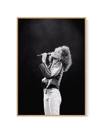 Framed black and white photograph of Whitney Houston singing into a microphone on stage, captured in a dynamic pose with her head tilted back and eyes closed, wearing a shimmering top and light-colored jeans, framed in natural oak with a white border.