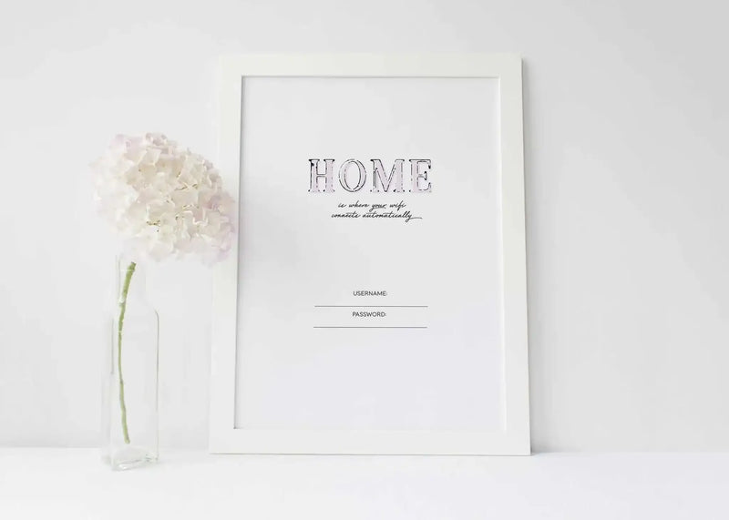 Framed art print featuring the word "HOME" in large, distressed black capital letters, with "is where your wife connects automatically" in a smaller, elegant script below it. The print also includes "USERNAME:" and "PASSWORD:" with lines for input, all on a clean white background. It is displayed in a simple white frame with a white border. A clear glass rectangular vase with a single white hydrangea bloom is positioned to the left of the print.