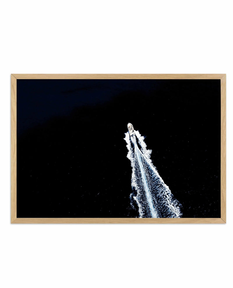 Photography art print featuring an aerial view of a white speedboat cutting through dark water, leaving a long, white, foamy wake behind it. The composition is dramatic with the dark water contrasting sharply with the bright white boat and wake, displayed in a natural oak frame with white border.