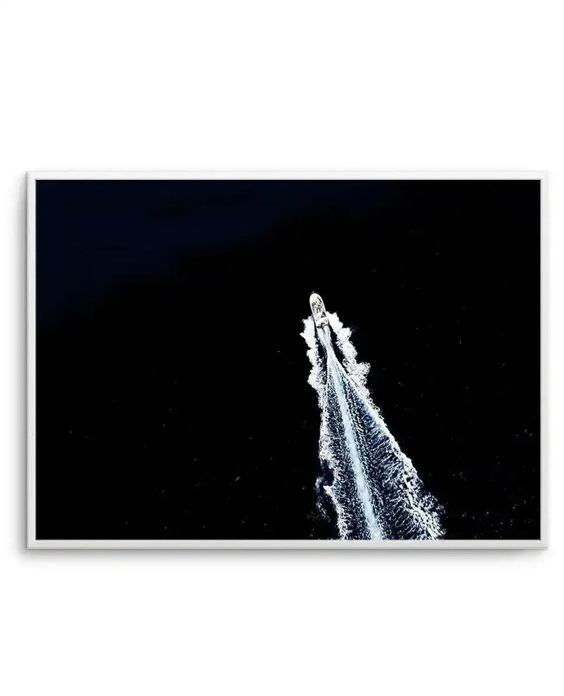 Framed art print featuring a dramatic overhead aerial photograph of a white speedboat cutting through dark navy water, creating a bright wake trail. The boat appears as a small bright form at the top of the composition with sweeping white spray patterns extending downward against the nearly black water. The image has high contrast between the luminous white vessel and wake against the deep dark background, creating a striking minimalist composition. The print is mounted in a white frame with a white border.