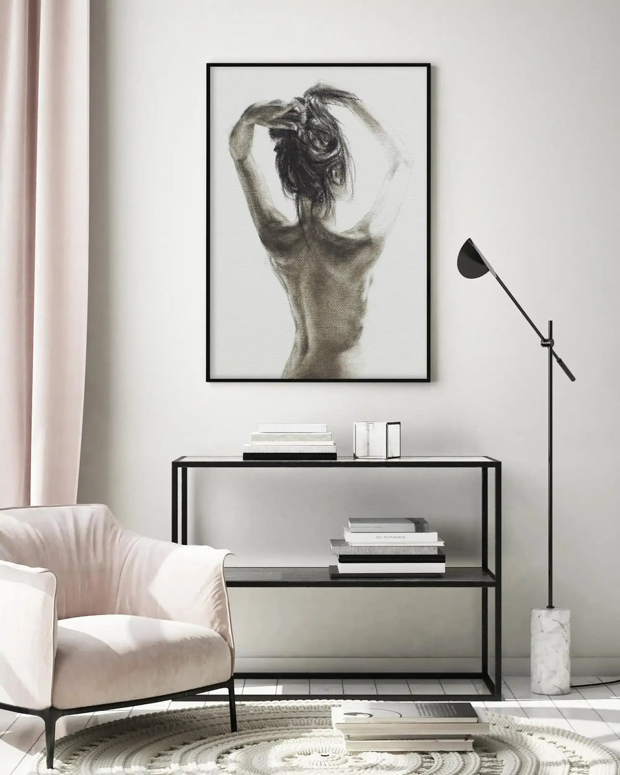 Lifestyle shot of a minimalist living room featuring a framed art print of a woman's back in charcoal. The print, with a black frame and white border, depicts a woman from the back, her arms raised as if tying her hair, rendered in charcoal with visible texture against a light background. The room includes a light pink armchair, a black metal console table with books and decorative objects, a cream textured rug, and a black floor lamp with a marble base.
