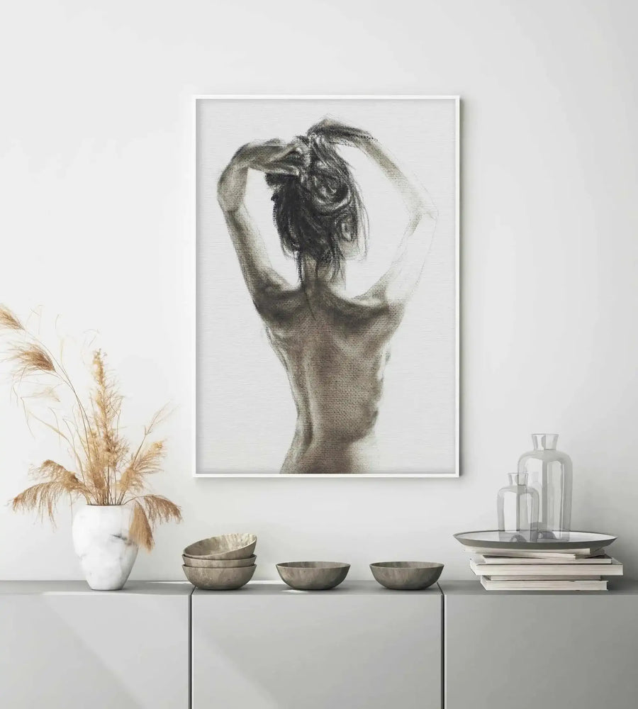 Lifestyle shot of a minimalist room featuring a framed art print of a woman's back in charcoal. The print, displayed in a white frame with a white border, shows a figure from the back, with arms raised and hands in her hair, rendered with visible charcoal strokes in shades of grey and black on a white background. Below the print, a light grey cabinet holds a white marble vase with dried pampas grass, a stack of grey bowls, and a tray with clear glass vases on a stack of books.