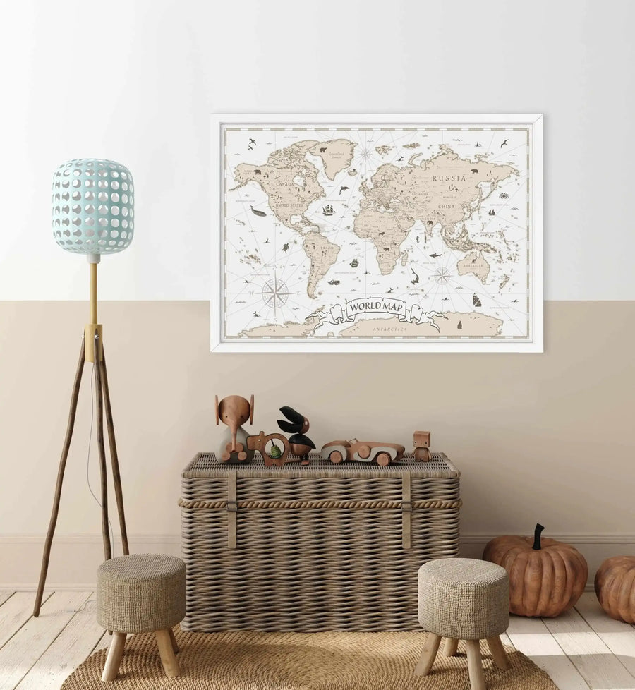 Lifestyle shot of a child's room featuring a framed world map art print above a woven toy chest. The map is a vintage-style illustration with countries in a light beige and oceans in off-white, featuring hand-drawn animals, ships, and a compass rose. The frame is white with a white border. The room has a two-tone wall, light wood floor, a light blue tripod lamp, two small woven stools, and decorative pumpkins.